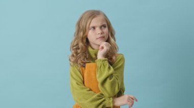 Sarı kıvırcık saçlı düşünceli genç kız bir şeyler düşünüyor ve mavi arka planda izole edilmiş bir fikir jesti yapıyor.