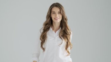 Ofis kıyafetleri içindeki güzel genç bir kadın ellerini stüdyodaki gri bir arka planda birleştirirken kendine güvenli bir poz veriyor.