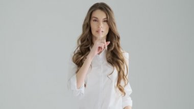 Sakin genç bir kadın, stüdyodaki gri arka planda izole edilmiş bir sessizlik jesti yapıyor.