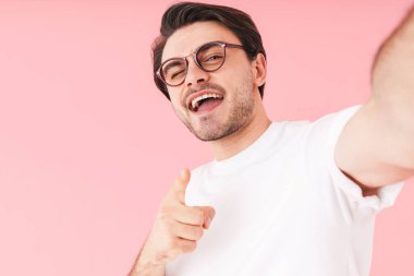 Beyaz gözlüklü, gözlüklü, kamerayı işaret eden ve pembe arka planda izole edilmiş selfie çeken bir adam resmi.