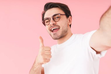 Kafkasyalı gözlüklü, gözlüklü, başparmağıyla hareket eden ve pembe arka planda izole edilmiş selfie çeken bir adam resmi.