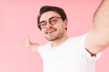 Gözlük takan, göz kırpan ve pembe arka planda izole edilmiş selfie çeken genç adamın görüntüsü.