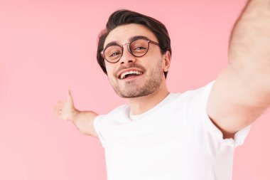 Gözlük takan, gülümseyen ve pembe arka planda izole edilmiş selfie çeken genç bir adam resmi.
