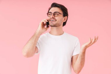 Gözlüklü, cep telefonuyla konuşan ve pembe arka planda izole edilmiş gülümseyen genç bir adam görüntüsü.