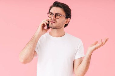 Kafası karışmış, gözlüklü, cep telefonuyla konuşan ve pembe arka planda izole edilmiş genç bir adam.