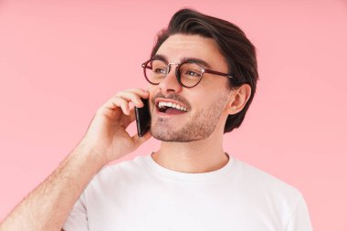 Gözlüklü, cep telefonuyla konuşan ve pembe arka planda izole edilmiş gülümseyen genç bir adam görüntüsü.