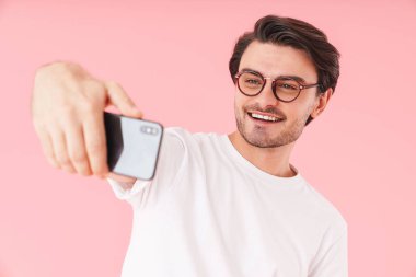 Gözlük takan, cep telefonuyla selfie çeken ve pembe arka planda izole bir şekilde gülümseyen yakışıklı bir adam.