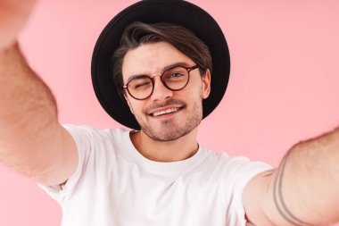 Gözlüklü ve şapkalı iyimser bir adamın pembe arka planda izole edilmiş selfie çekerken gülümsemesi.