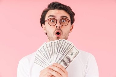 Gözlüklü, elinde nakit para olan ve pembe arka planda izole edilmiş kameraya bakan şaşırmış bir genç resmi.