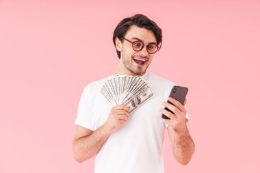 Gözlüklü, elinde nakit para olan ve pembe arka planda izole edilmiş cep telefonu kullanan genç bir adamın görüntüsü.