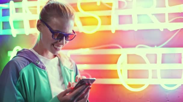 3Dメガネの美しい若い女性は 多色のネオンテキスト上のスマートフォンを使用しています — ストック動画