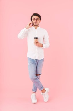 Tıraşsız, gözlüklü, kahve ısmarlayan ve pembe arka planda izole bir şekilde cep telefonuyla konuşan şaşırmış bir adamın görüntüsü.
