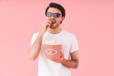 Üç boyutlu gözlük takan, patlamış mısır yiyen ve pembe arka planda izlenmiş bir film izleyen komik bir genç resmi.