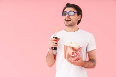 3 boyutlu gözlüklü esmer adamın elinde patlamış mısır ve soda şişesiyle pembe arka planda duran bir kova.