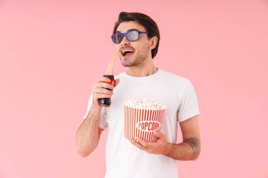 3 boyutlu gözlüklü, elinde patlamış mısır ve soda şişesiyle pembe arka planda duran heyecanlı bir adam resmi.