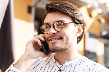 Kafede çalışırken cep telefonuyla konuşan, gözlüklü, gülümseyen beyaz bir adamın fotoğrafı.