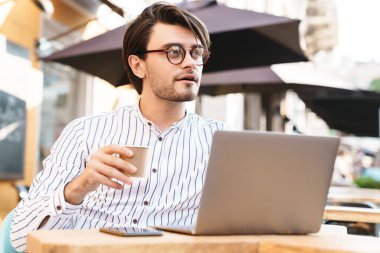 Kafkasyalı bir adamın dışarıda kahve içerken dizüstü bilgisayar ve cep telefonu kullanarak gözlük taktığını gösteren bir fotoğraf.