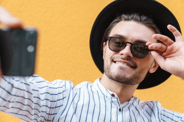 Güneş gözlüğü takan, siyah şapkalı, gülümseyen ve cep telefonunda selfie çeken genç bir adamın görüntüsü sarı arka planda izole edilmiş.