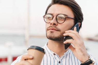 Gözlüklü genç adamın cep telefonuyla konuştuğunu ve iskelede yürürken kağıt bardaktan kahve içtiğini gösteren fotoğraf.