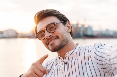 Gözlüklü, kamerayı işaret eden ve gezinti güvertesinde yürürken selfie çeken başarılı bir adamın fotoğrafı.