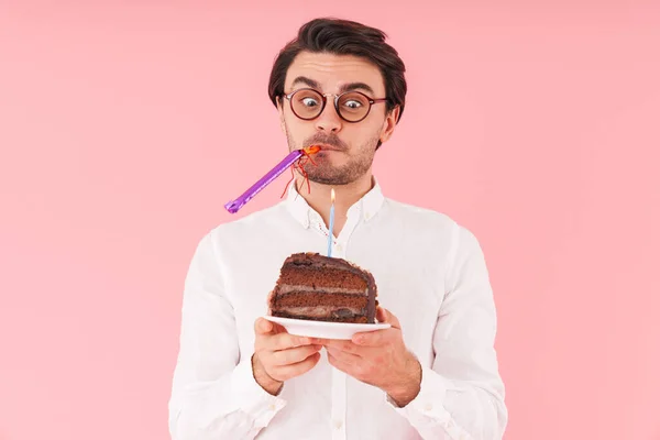 ピンクの背景に隔離されたキャンドルとチョコレートケーキを保持眼鏡をかけて若い驚きの男のイメージ — ストック写真