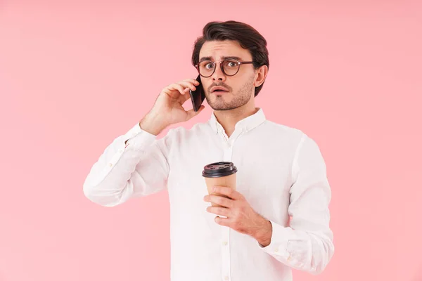 图片中的高加索人心烦意乱的男人戴着眼镜 一边喝着咖啡 一边在粉色背景下用孤立的手机交谈 — 图库照片