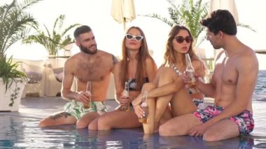 Dört gülümseyen çekici genç arkadaş havuz kenarında oturmuş bira içip sohbet ediyorlar.