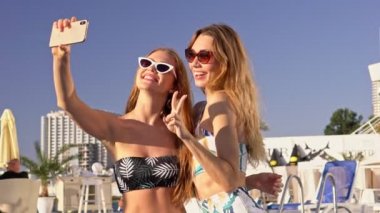 Mayolu ve güneş gözlüklü iki güzel kadın havuz başında selfie çekiyor ve eğleniyorlar.
