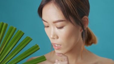 Çekici genç Asyalı bir kadın yeşil bir bitki yaprağı tutuyor ve mavi duvarın arkasından ona bakıyor.