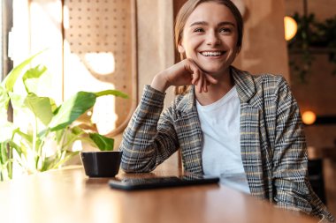 Kahve içen ve rahat bir kafede otururken cep telefonu kullanan mutlu genç bir kadının fotoğrafı.