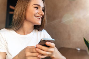 Neşeli genç bir kadının cep telefonu kullanıp rahat bir kafede otururken gülümsediği bir fotoğraf.