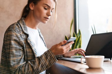 Bilgisayarda yazan ve rahat bir kafede otururken cep telefonu kullanan çok çekici bir kadının fotoğrafı.