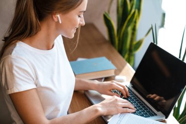 Gülümseyen güzel bir kadının, rahat bir kafede otururken dizüstü bilgisayar ve kulaklıklarla ders çalışmasının fotoğrafı.