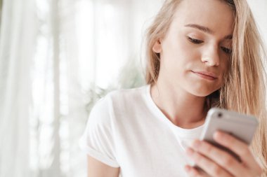 Sarı saçlı, güneşli odada akıllı telefondan yazı yazan hoş bir genç kadın resmi.