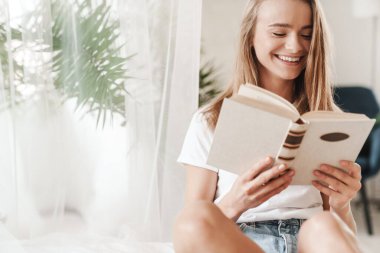 Güneşli bir odada yatakta oturmuş, sarışın saçlarıyla kitap okuyan, gülen genç bir kadın resmi.