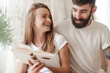 Evdeki parlak odada otururken gülümseyen ve kitap okuyan mutlu beyaz bir çift.