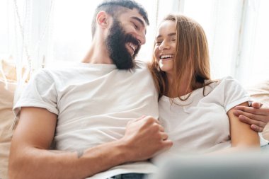 Romantik, gülen bir çiftin evde otururken dizüstü bilgisayar kullanması ve kucaklaşması.