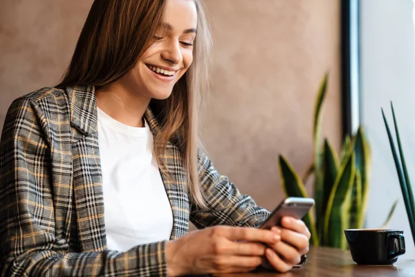 照片上的年轻女子坐在舒适的咖啡店里 一边喝咖啡一边用手机 — 图库照片