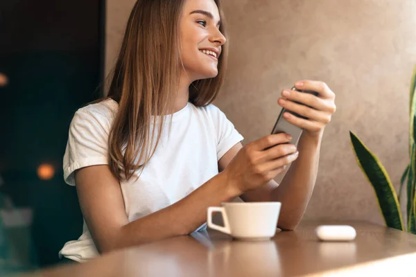 Fotografie Usměvavé Mladé Ženy Pití Kávy Pomocí Mobilního Telefonu Při — Stock fotografie