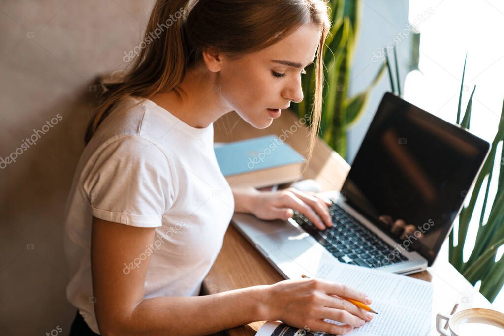 Photo of serious cute woman using laptop and studying with exercise book while sitting in cozy cafe