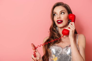 Pembe duvar arkasında izole bir şekilde telefonla konuşan parlak pullu elbiseli, gülümseyen genç bir kadın görüntüsü.