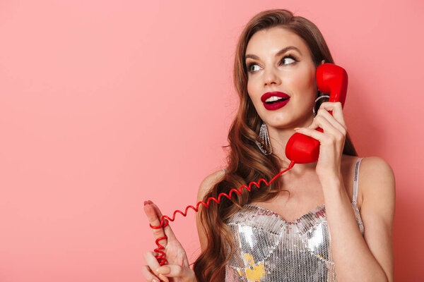 Изображение молодой улыбающейся женщины в ярких блестках платье изолированы на розовом фоне стены говорят по телефону
.