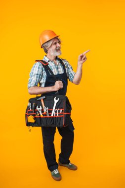 Kasklı yaşlı, gri saçlı, sakallı bir inşaatçının fotoğrafı. Sarı duvar arkasına izole edilmiş bir şekilde poz veriyor. Elinde çanta ve ekipman gereçleri var..