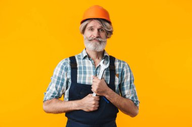 Olumlu iyimser, yaşlı, gri sakallı, miğferli adam görüntüsü. Sarı duvar arkasında ingiliz anahtarı tutuyormuş gibi duruyor..