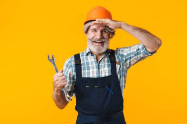Kasklı, mutlu, gri saçlı, sakallı bir adam resmi. Sarı duvar arkasında ingiliz anahtarı tutuyormuş gibi duruyor..