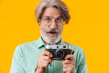 Şok olmuş gri saçlı fotoğrafçının görüntüsü. Sarı duvar arkasında kamerayla poz veriyor..