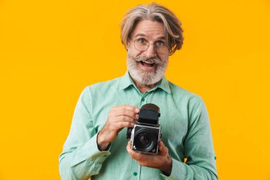 Sarı duvar arkasında kamerayla izole edilmiş mutlu, gri saçlı fotoğrafçı görüntüsü..