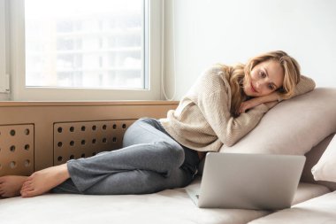 Evde dizüstü bilgisayar kullanan çok tatlı ve güzel bir genç kadının görüntüsü kanepede yatıyor..