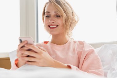 Evinde, battaniyenin altında cep telefonu kullanan neşeli, mutlu bir genç kadının görüntüsü..