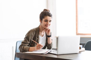 Dizüstü bilgisayar kullanan genç ve güzel bir kadının görüntüsü. Kapalı alanda not defteri ile notlar yazıyor..